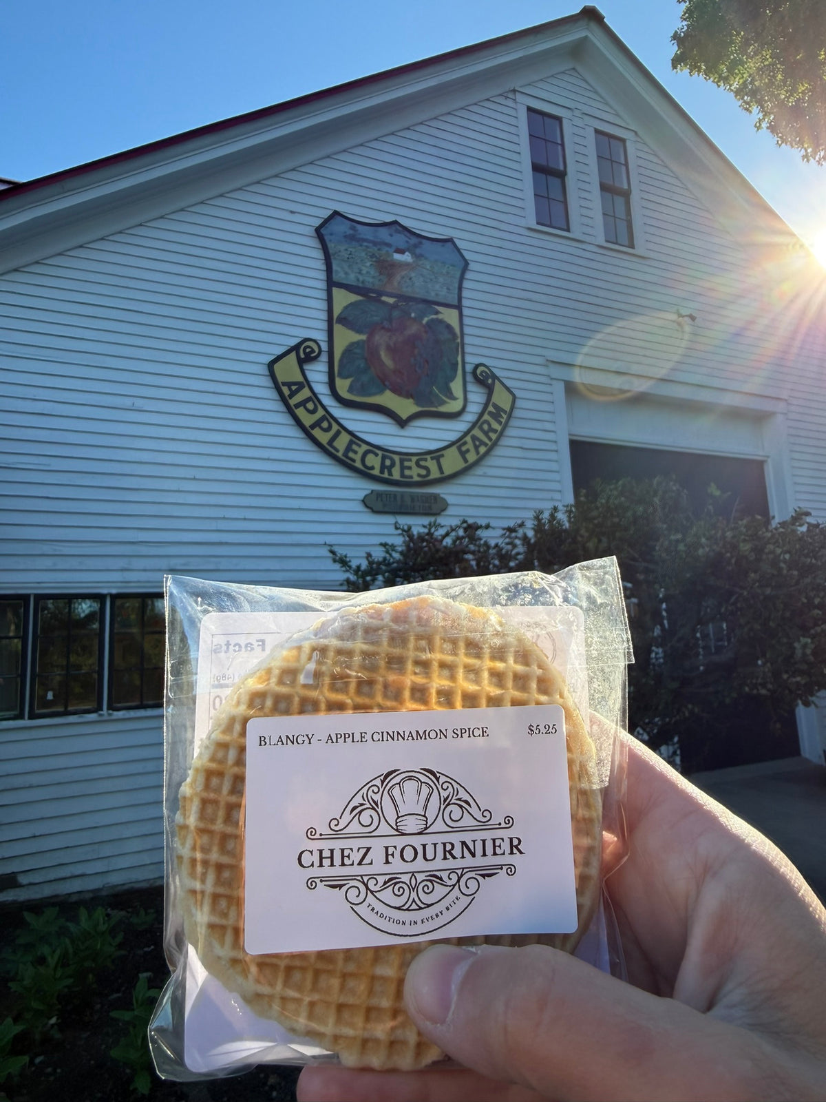 A hand holds a packaged round waffle in front of a white farmhouse with a crest sign reading AppleGrest Farm. The package label says Chez Fournier and the waffle flavor is Blangy - Apple Cinnamon Spice.