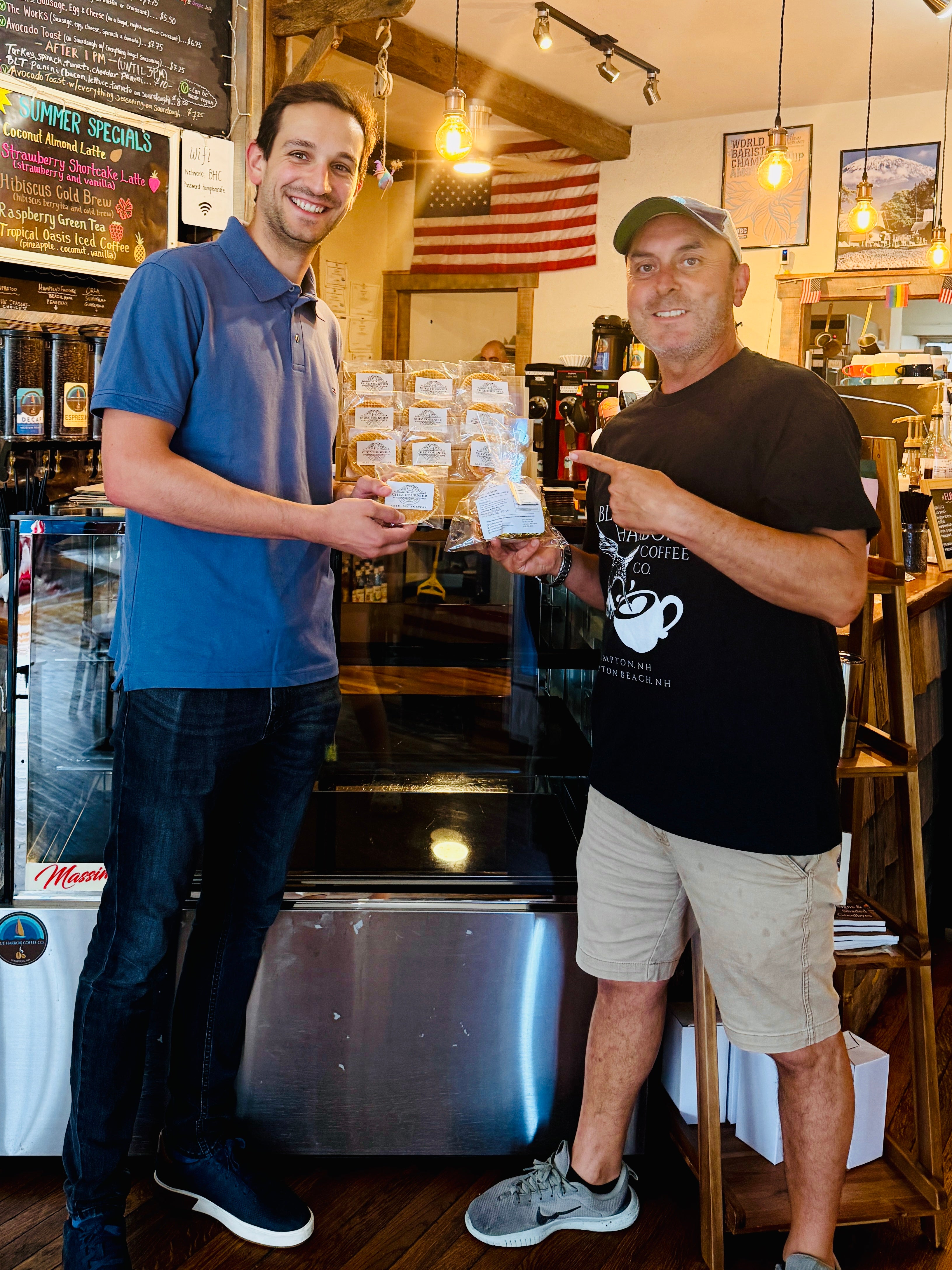 Two men standing inside Blue Harbor coffee shop, each holding a clear plastic bag filled with small packaged waffles. The shop has wood ceilings, hanging lights, a chalkboard menu, an American flag, and a stainless steel counter behind them.